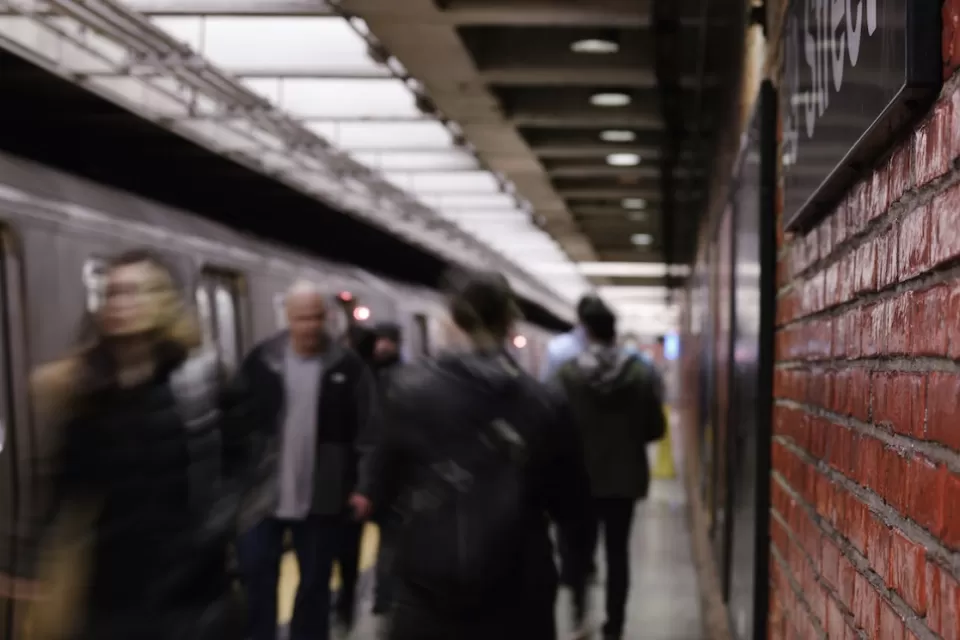 Busy New York City subway platform with blurred people walking in and out of trains and close-up on red brick wall on right.
